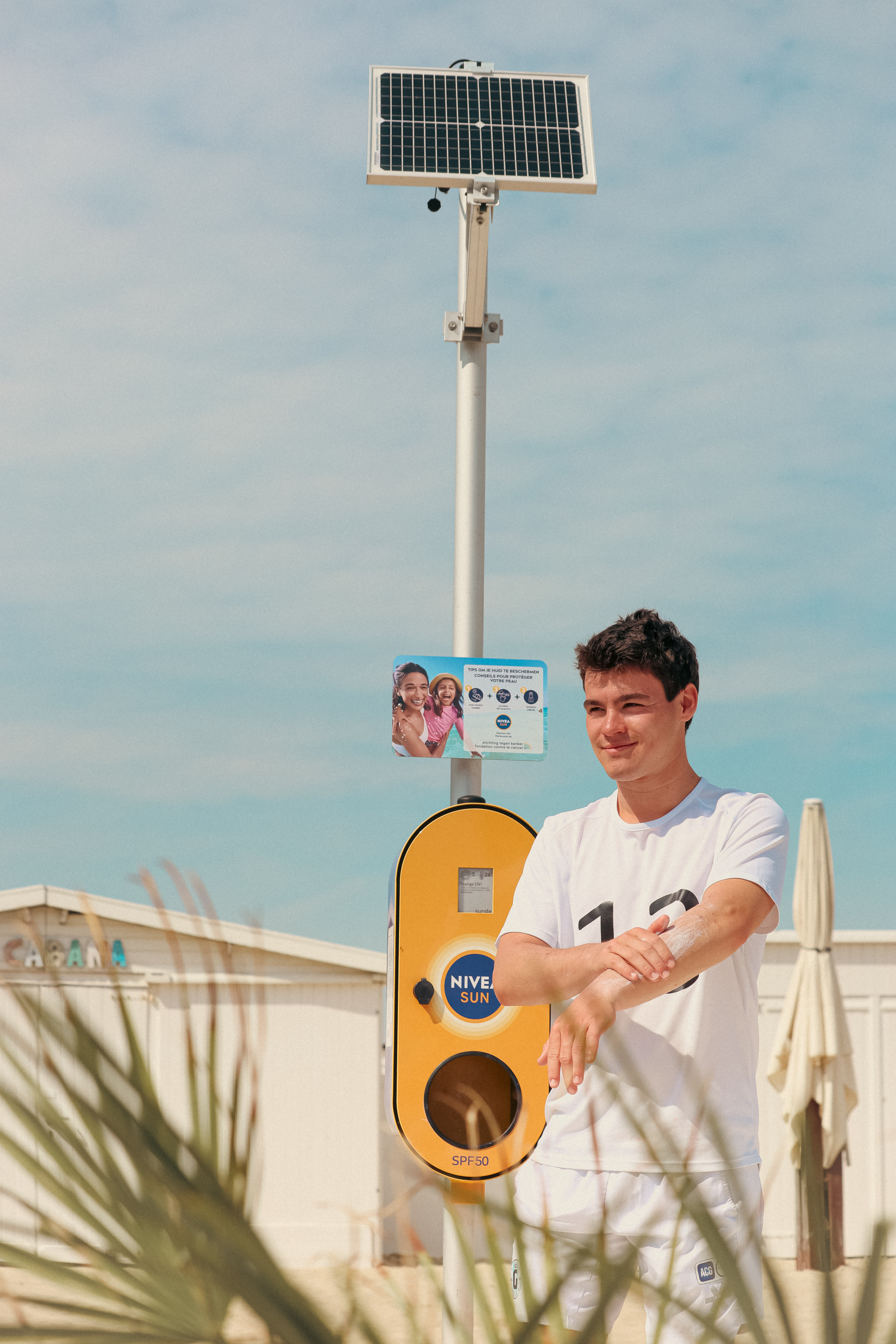 A young man applies sunscreen on his arm next to a SMOTspot: a smart solar-powered sunscreen dispenser placed on the beach. 
