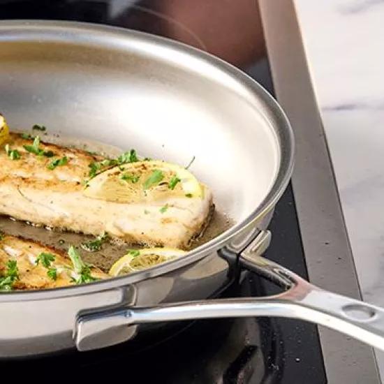 A stainless steel pan with fried fish fillets, garnished with lemon slices and parsley, on an induction cooktop. The pan has a nonstick texture as a PFAS-alternative.