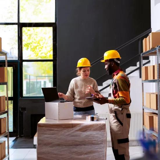 Deux employés d'entrepôt portant des casques discutent des processus de travail près d'un ordinateur portable, entourés d'étagères de boîtes, illustrant l'efficacité dans la réduction du Work-in-Progress (WIP).