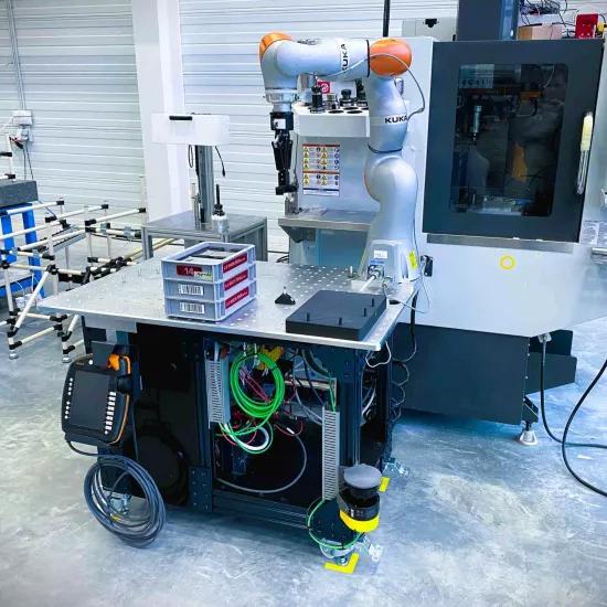 A mobile cobot assistant on a wheeled platform in an industrial environment, working next to a CNC machine. The cobot, branded KUKA, is designed to automate repetitive tasks in manufacturing. The setup includes cables, a control panel, and storage bins for small parts.