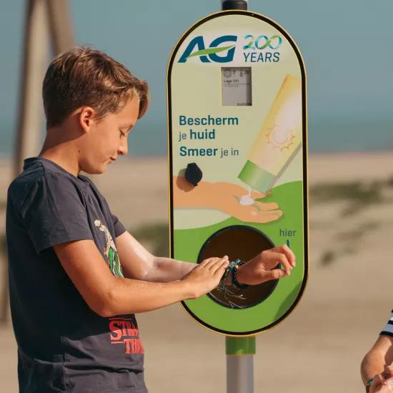 Two children apply sunscreen at a dispenser on the beach.