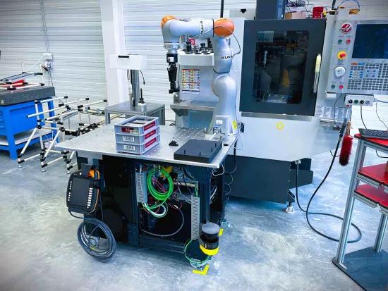 A mobile cobot assistant on a wheeled platform in an industrial environment, working next to a CNC machine. The cobot, branded KUKA, is designed to automate repetitive tasks in manufacturing. The setup includes cables, a control panel, and storage bins for small parts.