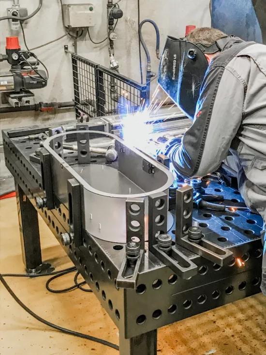 A metal worker welds a metal frame to a worktable using protective equipment and a welding hood.