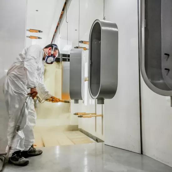 An employee in protective clothing applies powder coating to metal enclosures in a spray booth.