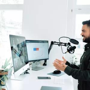 Man at standing desk webcasting in front of a professional microphone set and two large computer screens
